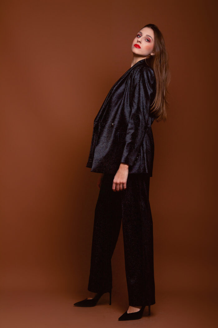 Devojka nosi crni LETITIA blejzer sa sjajnim šljokicama. Stoji ispred tamno crvene pozadine.