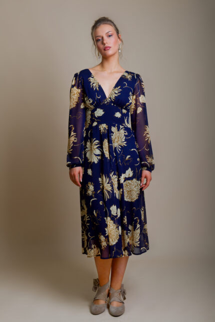 The girl is wearing a navy MARIA midi dress with beige flowers. She stands in front of a beige background and has ballet flats in the same color.