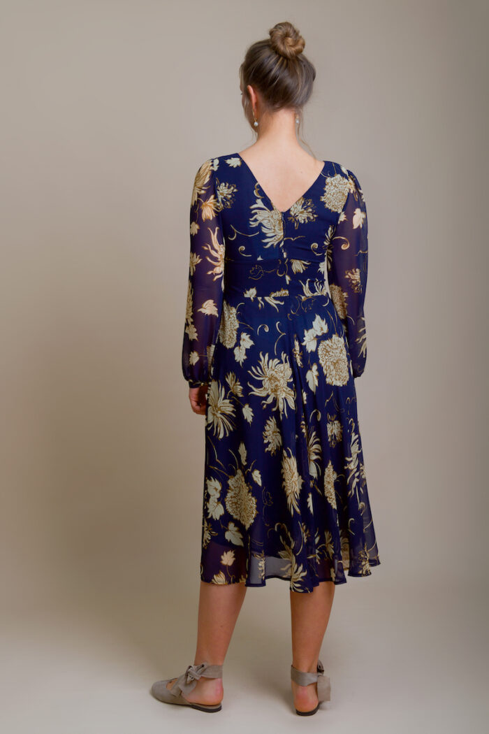 The girl is wearing a navy MARIA midi dress with beige flowers. She is standing with her back in front of a beige background and has ballet flats in the same color.