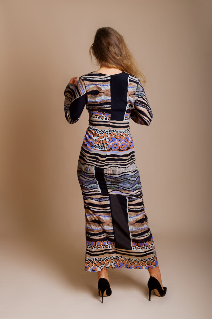 Devojka nosi UNA haljinu od dezenirane svile u animal printu, sa mnoštvom dizajnerskih detalja. Devojka nosi crne cipele sa visokim potpeticama i stoji ispred bež pozadine.