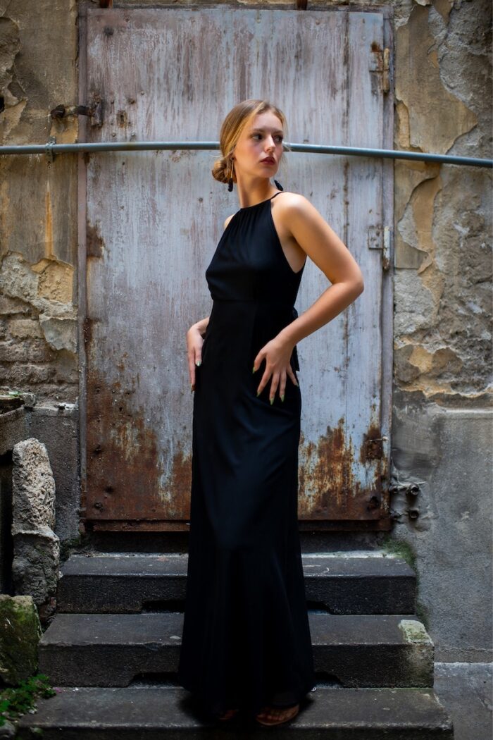 Plava devojka nosi dugu crnu svilenu HELENA haljinu i stoji ispred rustične pozadine.