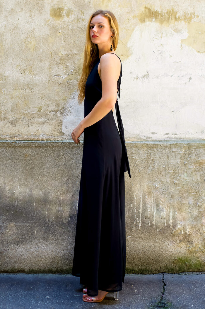 A blonde girl wears a long black silk HELENA dress and stands in front of a rustic beige background.
