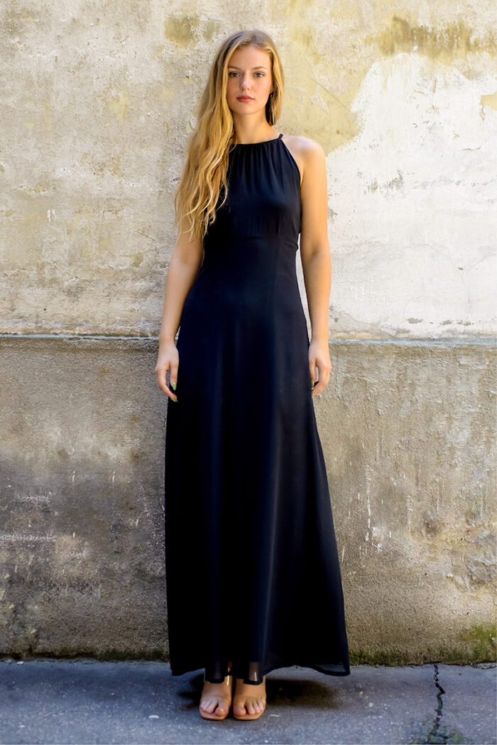 A blonde girl wears a long black silk HELENA dress and stands in front of a rustic beige background.
