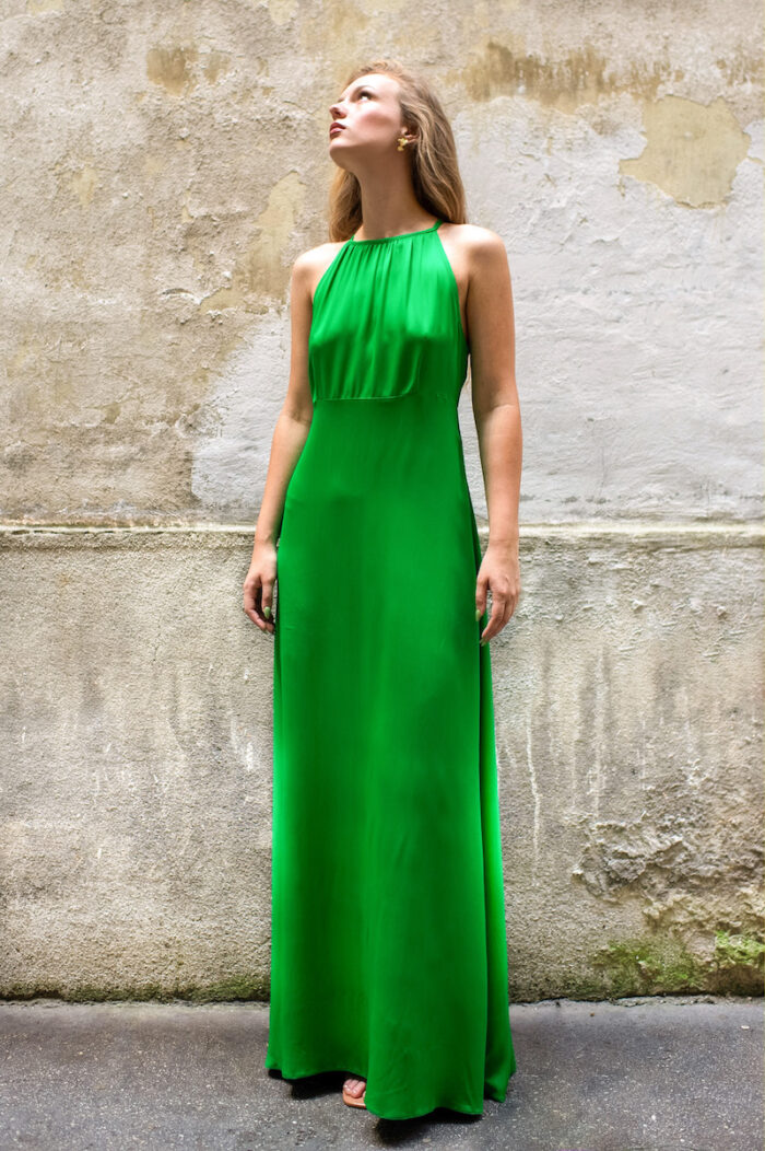 The blonde girl wears a long green HELENA viscose dress and stands in front of a beige rustic wall.