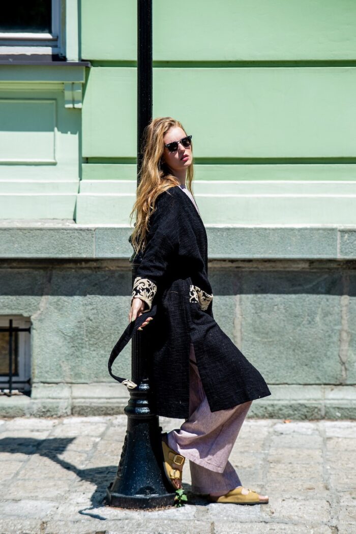 Devojka nosi crni pamučni NOA kardigan i stoji na ulici ispred zelenog zida.