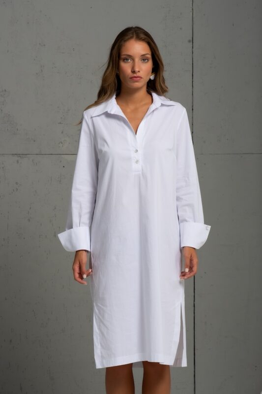 Brineta nosi belu pamučnu košulju midi dužine i stoji ispred sive pozadine.