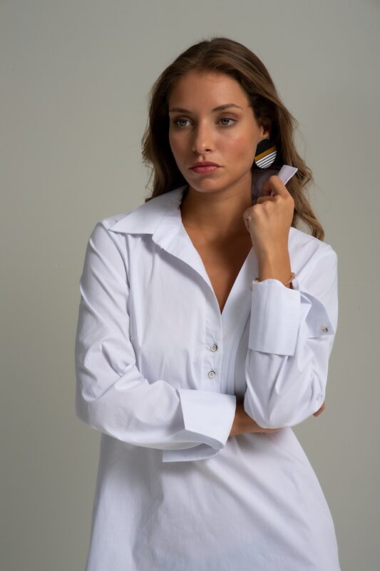 Brunette is wearing a white cotton shirt of midi length and standing in front of a gray background.
