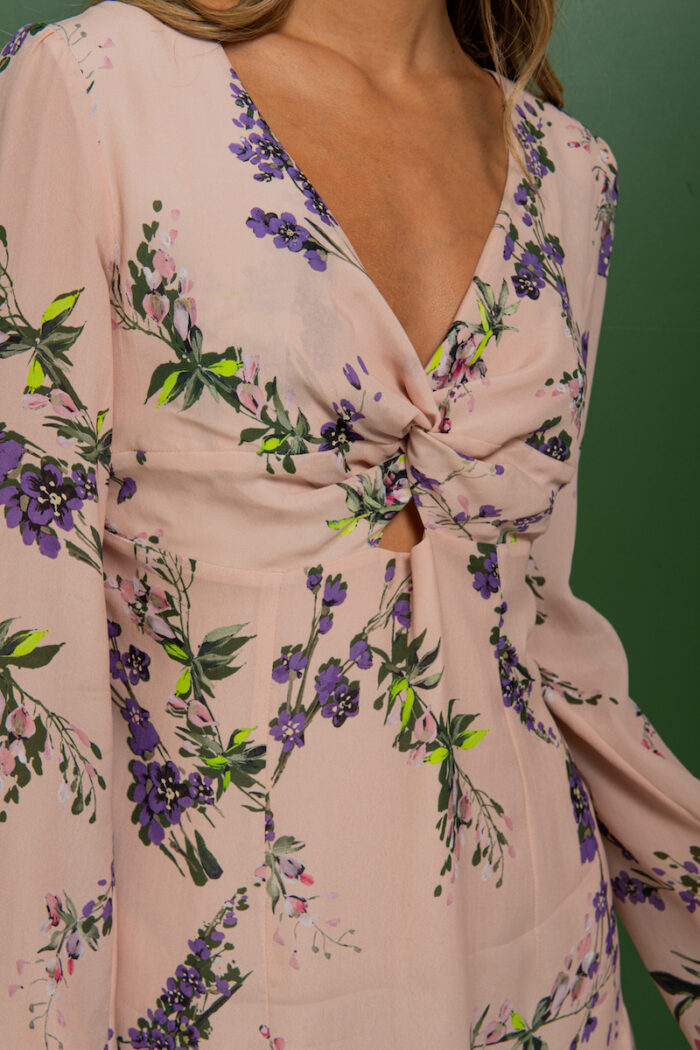 The brunette is wearing a short beige silk dress with a floral print and standing in front of a green background.