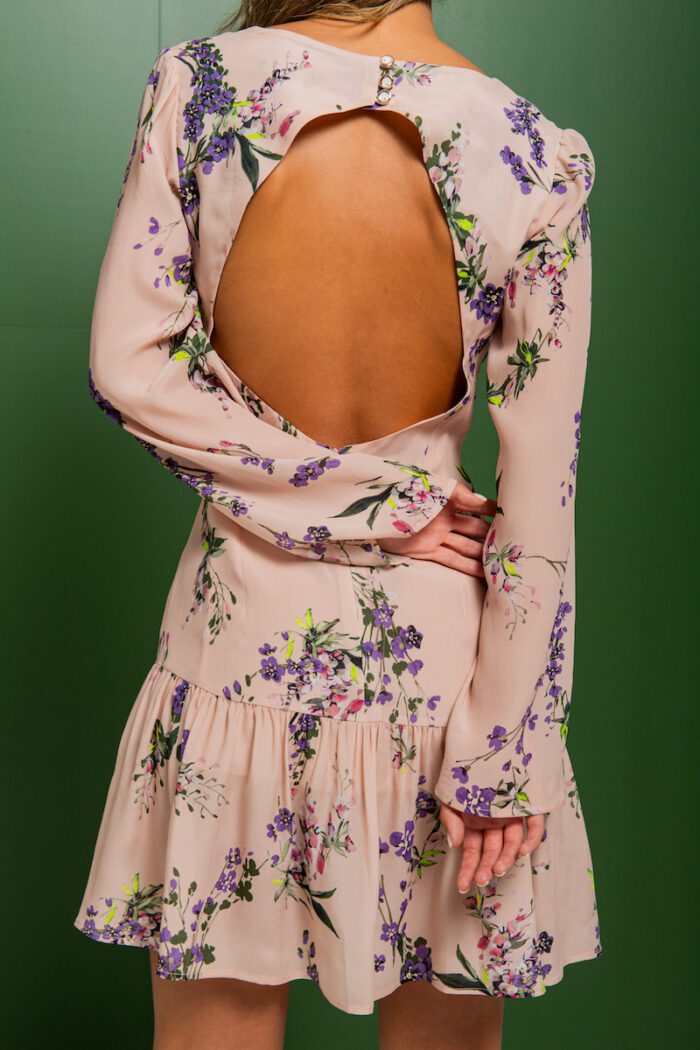 Brineta nosi kratku bež haljinu od svile sa floralnim printom i stoji ispred zelene pozadine.
