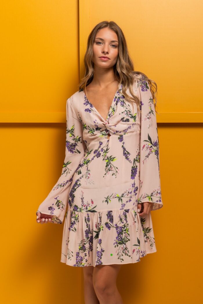 The brunette is wearing a short beige silk dress with a floral print and standing in front of a yellow background.