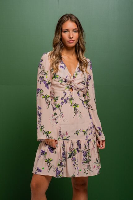 The brunette is wearing a short beige silk dress with a floral print and standing in front of a green background.