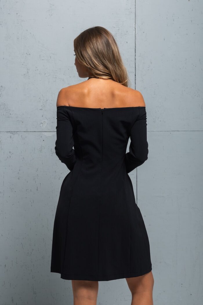 Brineta nosi malu crnu haljinu sa dugim rukavima i Bardo vratnim izrezom. Stoji ispred sivog zida.