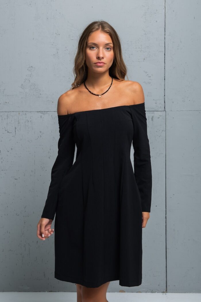The brunette is wearing a little black dress with long sleeves and a Bardot neckline. She is standing in front of a gray wall.