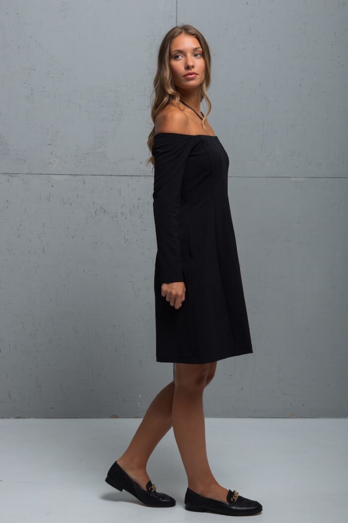 The brunette is wearing a little black dress with long sleeves and a Bardot neckline. She is standing in front of a gray wall.