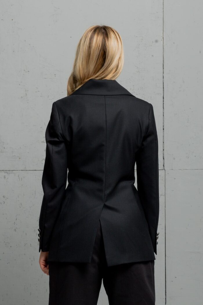 Plava devojka nosi klasični crni sako od hladne vune sa dvorednim kopčanjem i stoji ispred sivog zida.