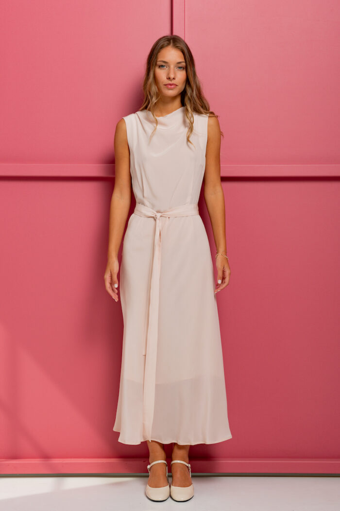 The girl is wearing a midi silk dress in powder pink color without sleeves, standing in front of a pink background.