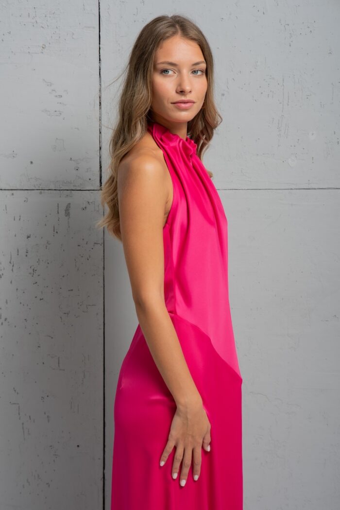 Brunette is wearing a long pink satin SIENA dress and standing in front of a gray wall.