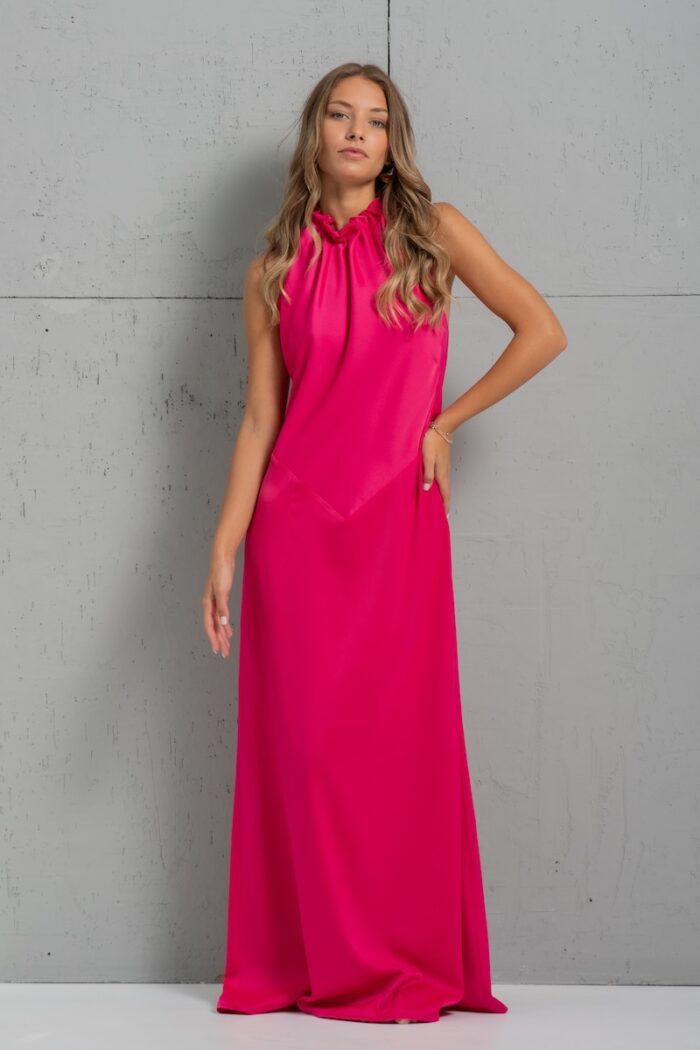 Brunette is wearing a long pink satin SIENA dress and standing in front of a gray wall.