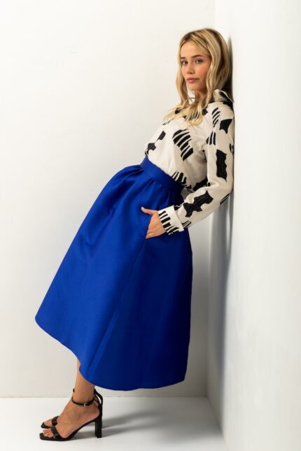 The blonde girl is wearing a blue BARBARA skirt and a white and black silk blouse, standing in front of a white wall.