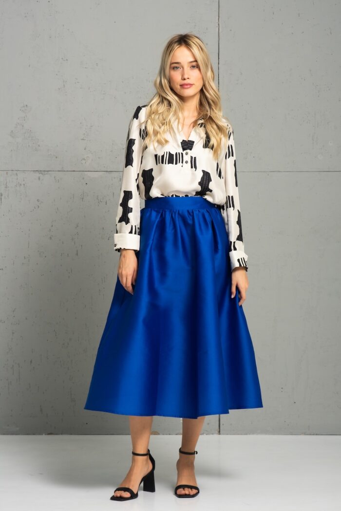 The blonde girl is wearing a blue BARBARA skirt and a white and black silk shirt and is standing in front of a white wall.