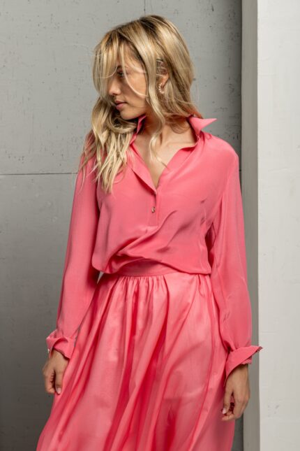 The blonde girl is wearing a light pink BELA silk shirt and standing in front of a gray wall.