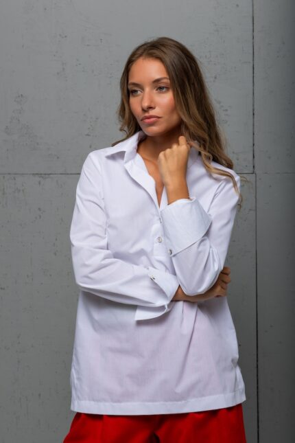 The brunette is wearing a white cotton BELA shirt with red shorts and standing in front of a gray wall.