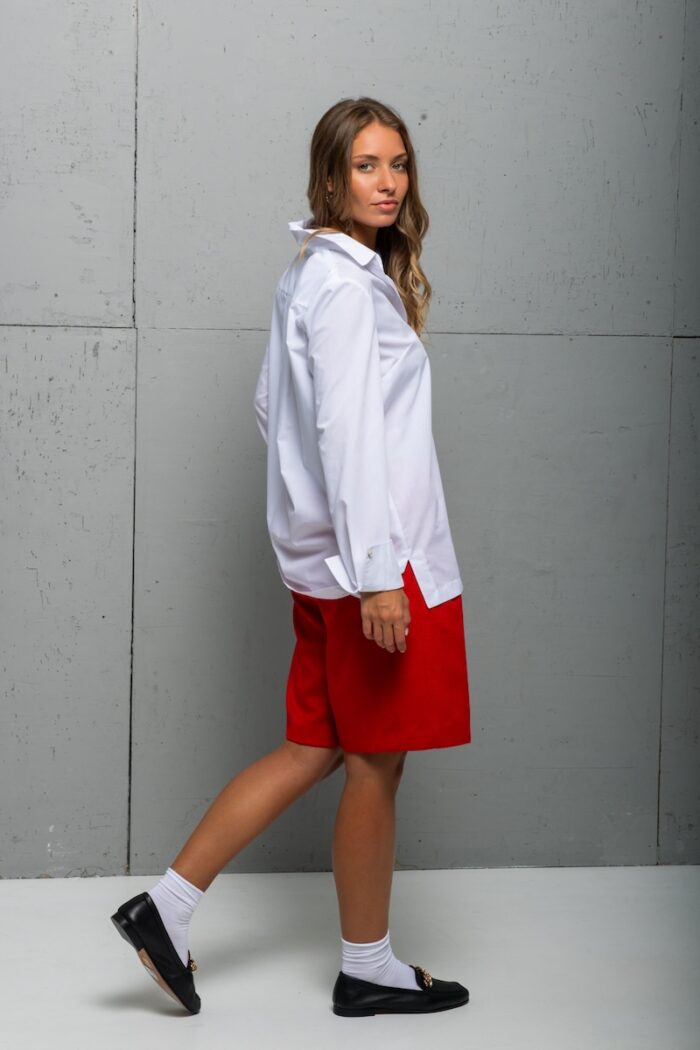 The brunette is wearing a white cotton BELA shirt with red shorts and standing in front of a gray wall