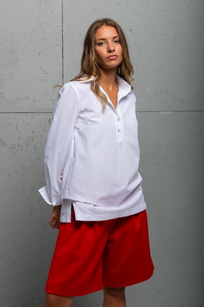 Brineta nosi belu pamučnu BELA košulju sa crvenim šortsem i stoji ispred sivog zida.