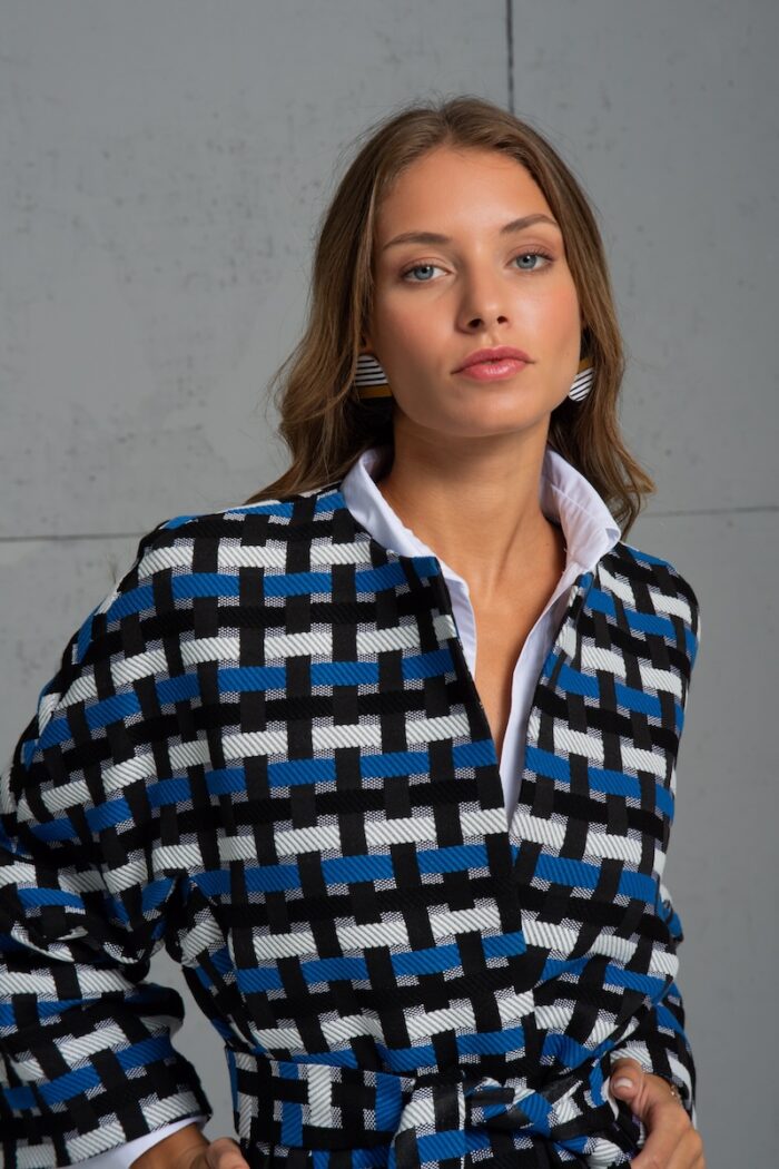 Brineta nosi GABRIELA kaput na vezivanje u plavo-crno-beloj kombinaciji boja i stoji ispred sive pozadine.