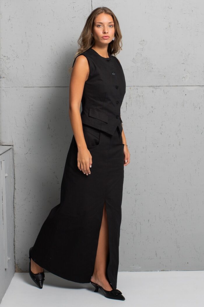 The girl is wearing a long skirt made of black twill with a slit at the front. She is standing in front of a gray wall.