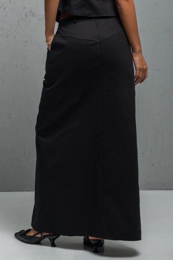 The girl is wearing a long skirt made of black twill with a slit at the front. She is standing in front of a gray wall.