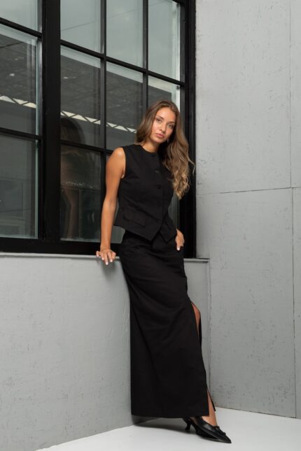 Devojka nosi dugu suknju od crnog kepera sa šlicem na prednjem delu. Stoji ispred sivog zida.