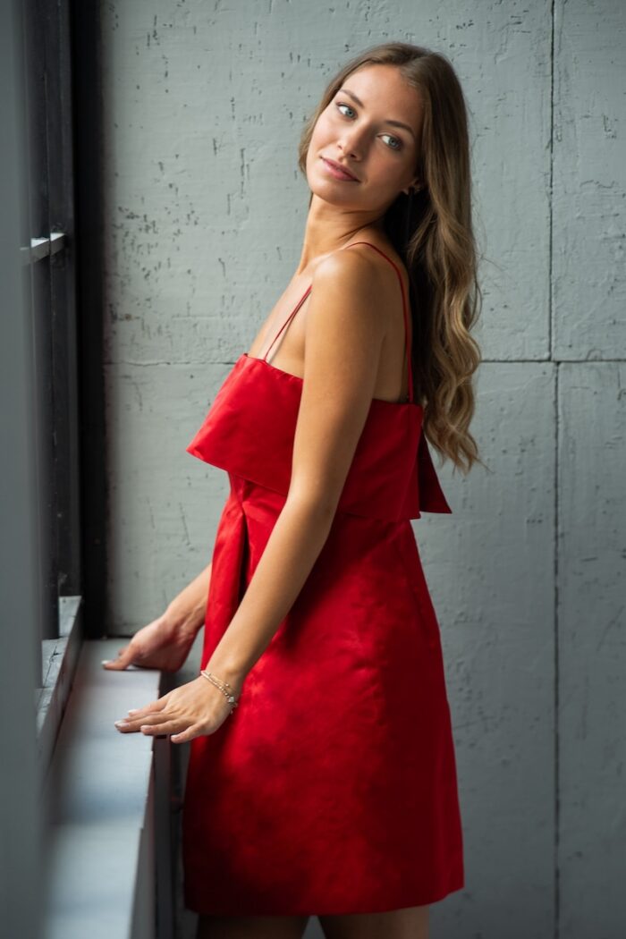 Devojka nosi kratku MADLENA haljina od satenskog materijala u tamno-crvenoj boji.
