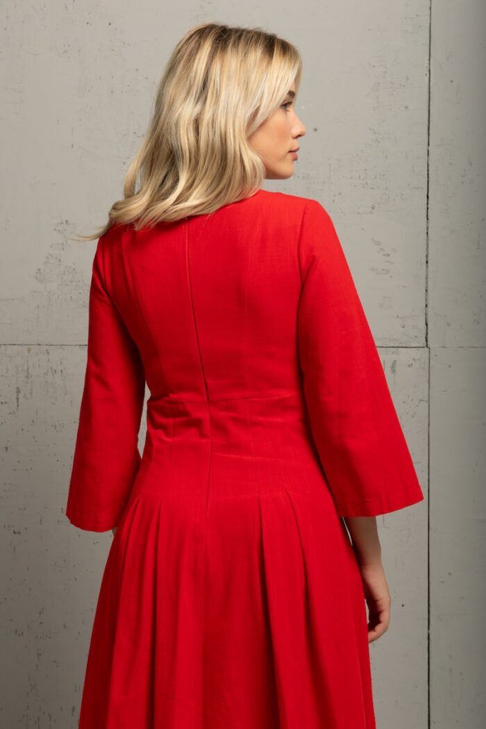 Plava devojka nosi crvenu midi MAXIMA haljinu i stoji ispred sivog zida.