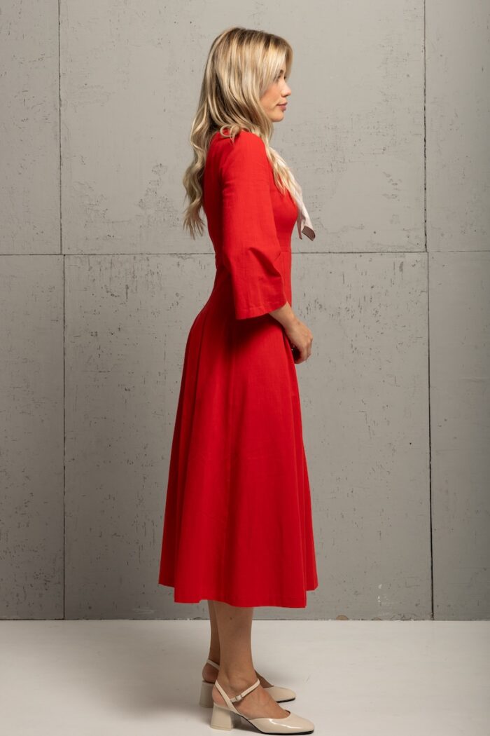 A blonde girl wears a red midi MAXIMA dress and stands in front of a gray wall.