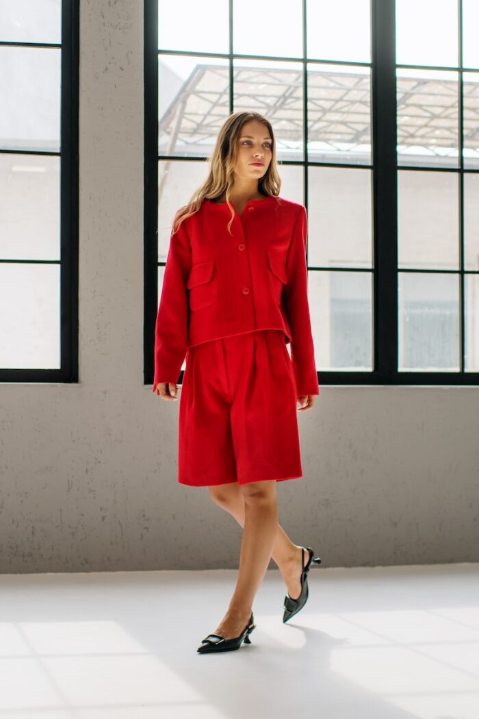 The girl wears a red RUBY jacket and red MINJA shorts with wide legs and stands in front of a gray wall.