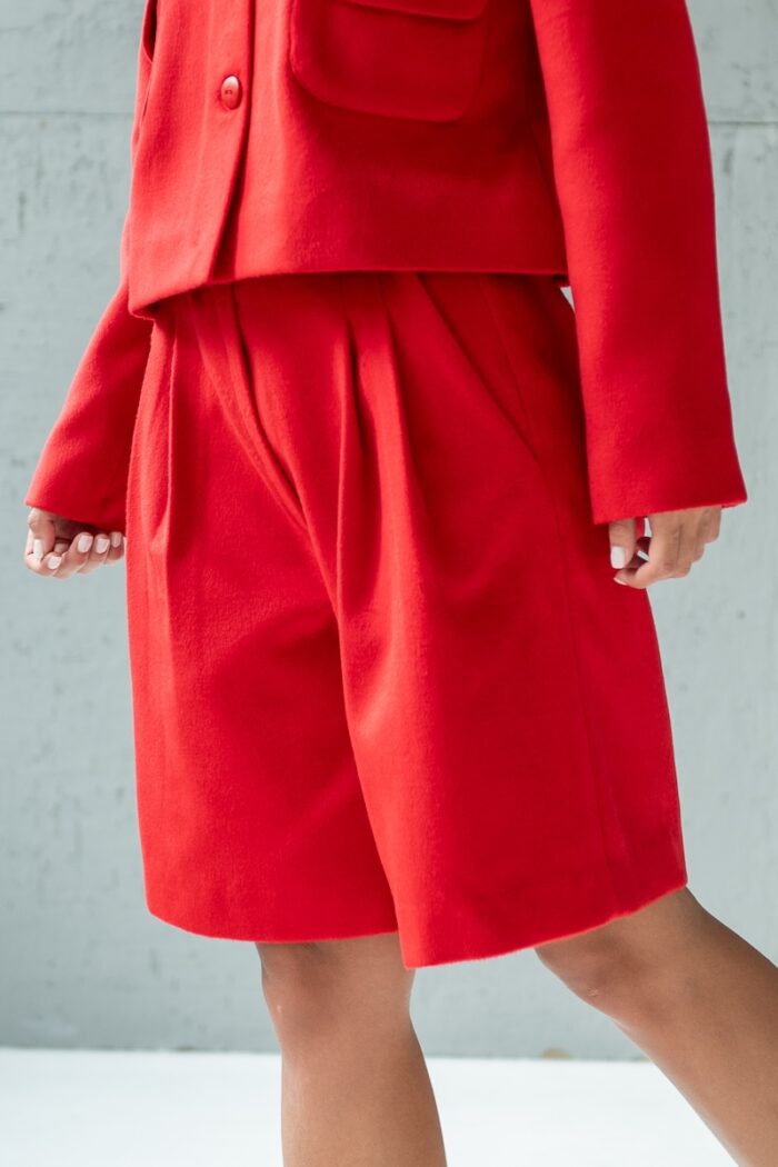 Devojka nosi crveni MINJA šorts sa širokim nogavicama i stoji ispred sivog zida.