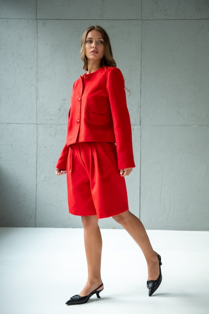 Devojka nosi crveni RUBY sako i crveni MINJA šorts sa širokim nogavicama i stoji ispred sivog zida.
