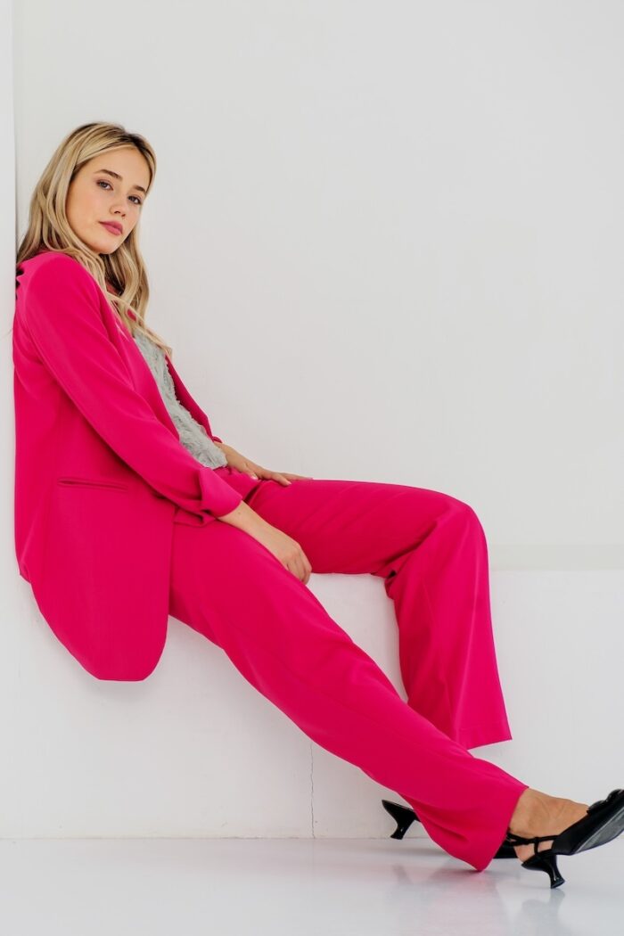 The blonde girl is wearing a pink set with a blazer and SONJA pants, sitting in a white room.