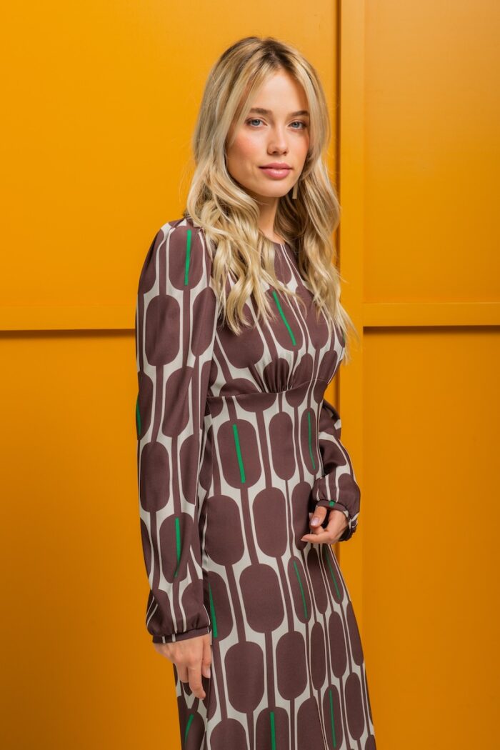 The blonde girl is wearing a silk midi TEONA dress with a brown-beige pattern and standing in front of a yellow wall.