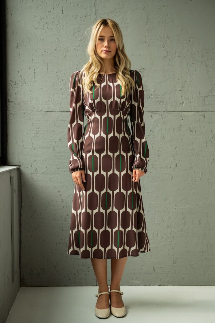 The blonde girl is wearing a silk midi TEONA dress with a brown-beige pattern and standing in front of a grey wall.