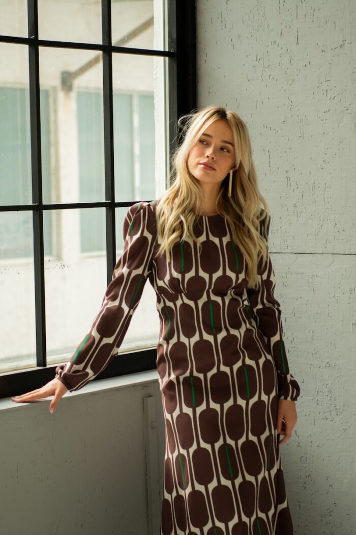 The blonde girl is wearing a silk midi TEONA dress with a brown-beige pattern and standing in front of a window and a grey wall.