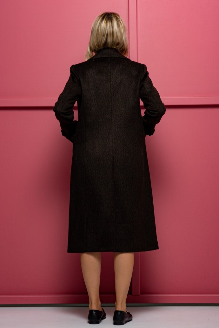 The blonde girl is wearing a woolen XENIA coat in brown with yellow buttons. She is standing in front of a pink background.