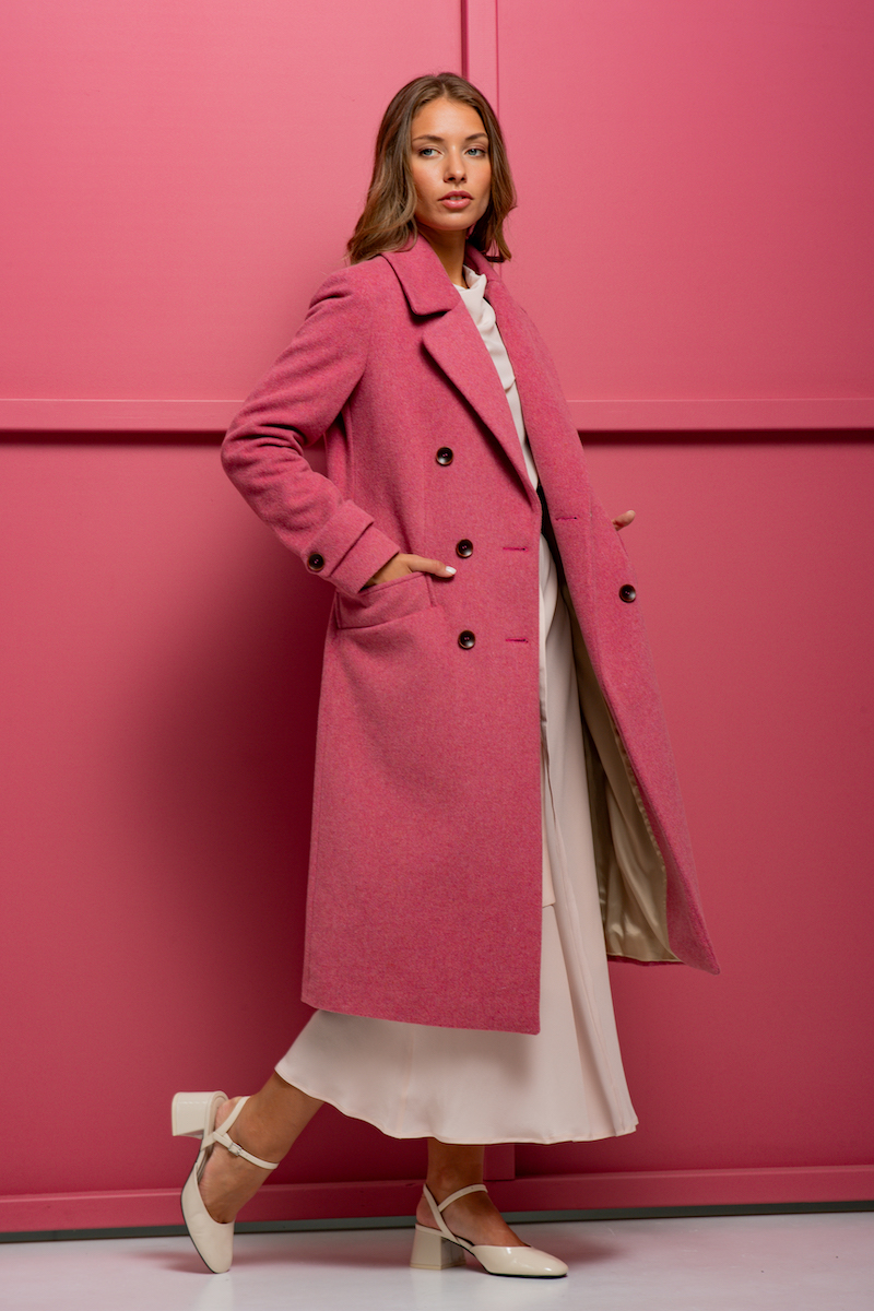 The brunette wears a striking pink XENIA coat and stands in front of a pink background.