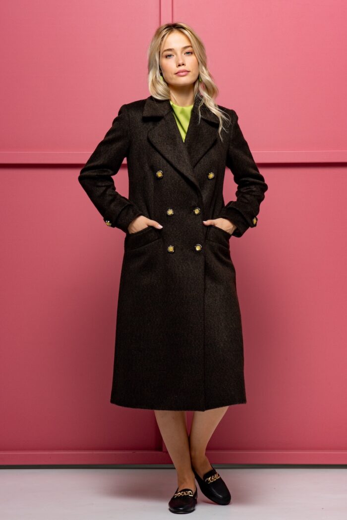The blonde girl is wearing a woolen XENIA coat in brown with yellow buttons. She is standing in front of a pink background.