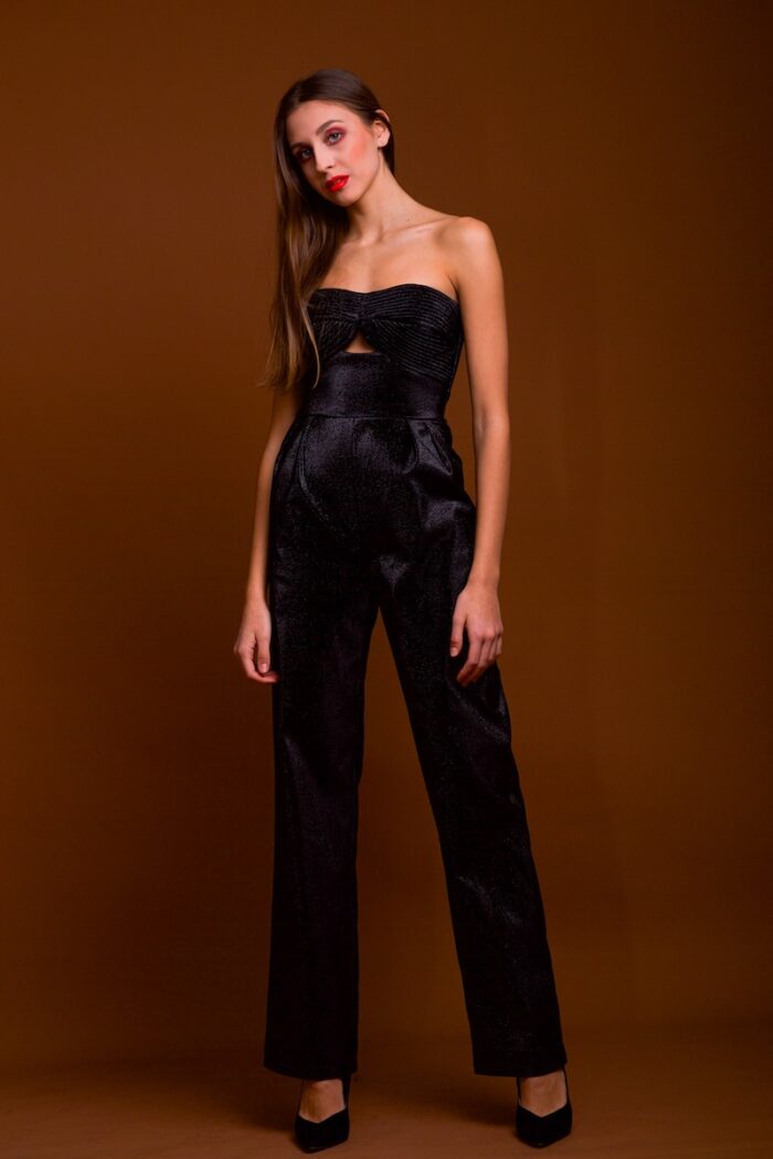 The girl wears a shimmering black jumpsuit with a prominent corset. She was photographed in front of a dark red background.