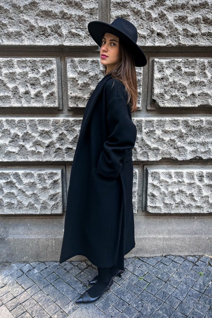 Devojka nosi crni kaput na vezivanje sa crnim šeširom i crnim cipelama. Slikana je ispred sivog zida.