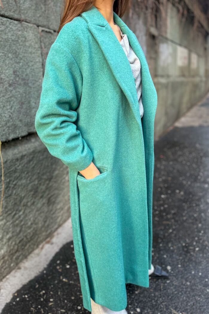 Devojka nosi zeleni CHARLOTTE kaput na vezivanje i drži ruke u džepovima.