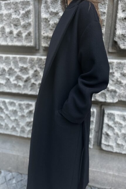 The girl is wearing a black tie-up coat with a black hat and black shoes. She is pictured in front of a gray wall.