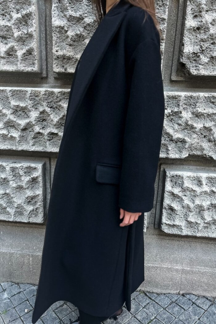 Devojka nosi crni kaput na vezivanje. Slikana je ispred sivog zida.